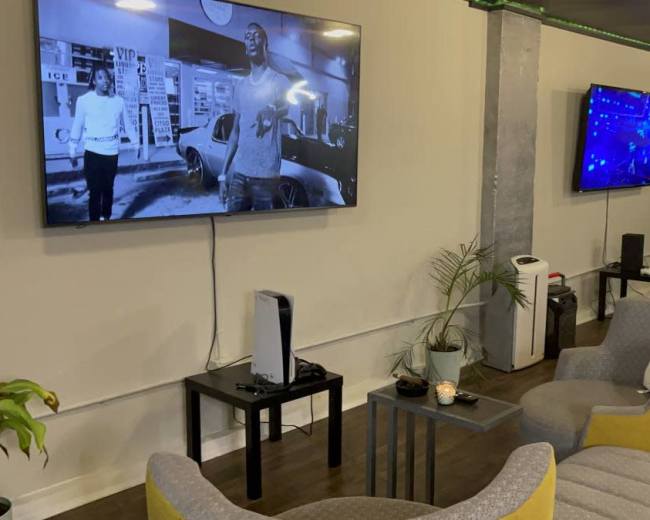 The room features two large wall-mounted TVs displaying different content, surrounded by modern seating and plants.