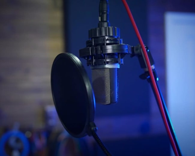 A microphone is positioned on a stand with a pop filter in a dimly lit recording studio.