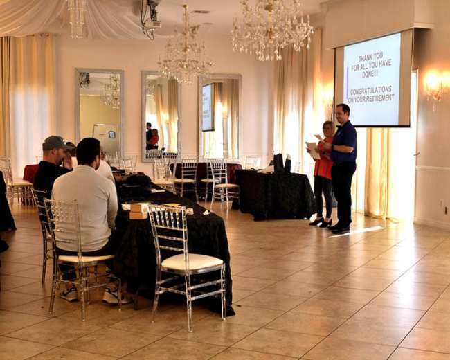 A man and a woman stand in front of a screen displaying a retirement message in a decorated hall with tables and chairs arranged for an event.