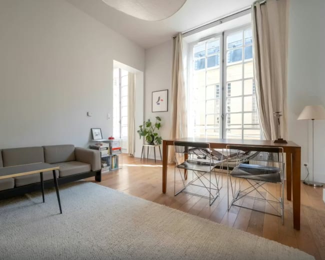 The image shows a minimalistic living space with a grey sofa, a wooden table with metal chairs, and large windows allowing natural light to illuminate the room.