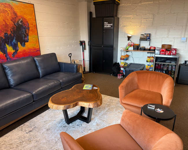 The image shows a cozy lounge area with a dark blue sofa, two orange armchairs, a wooden coffee table, and a small food and drink station in the background.