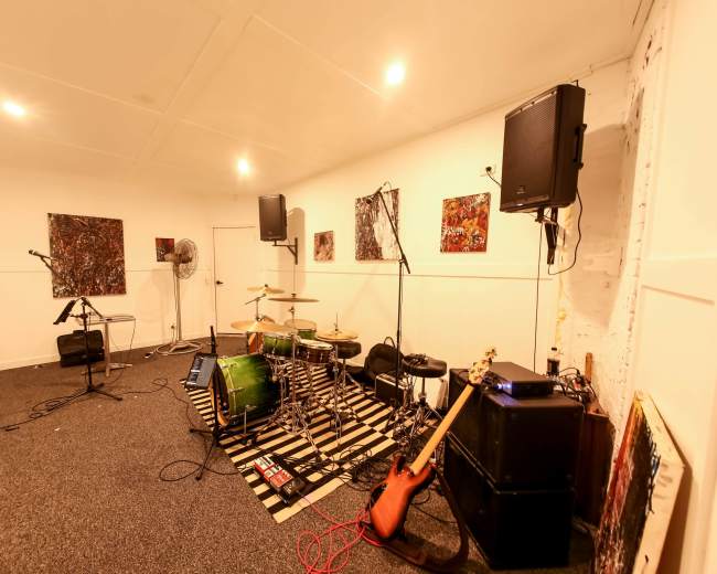 The image shows a music rehearsal space with a drum set, electric guitar, amplifiers, and sound equipment arranged along the walls.