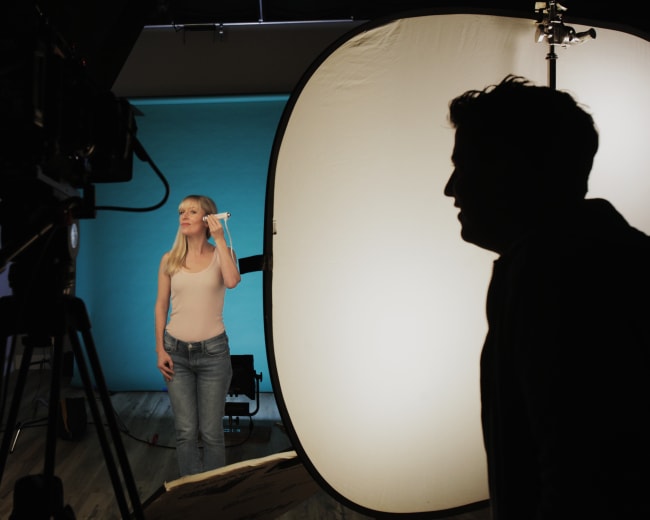 A model poses in front of a backdrop while being photographed by a crew using professional lighting equipment.