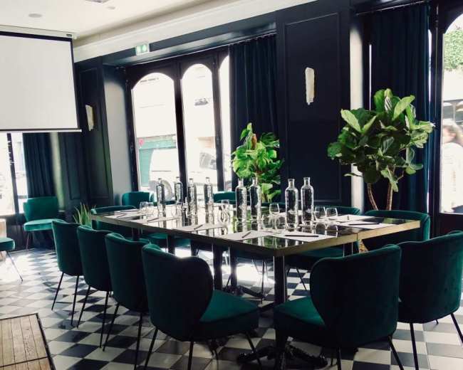 A conference room is set up with a long table surrounded by green chairs, adorned with plants, and featuring a projector screen and a checkered floor.