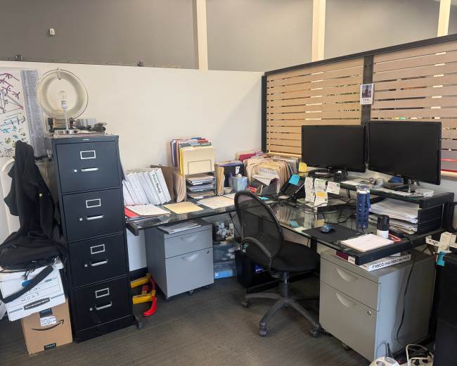 The image shows a cluttered office workspace with two computer monitors, filing cabinets, and stacks of paper on a glass desk.