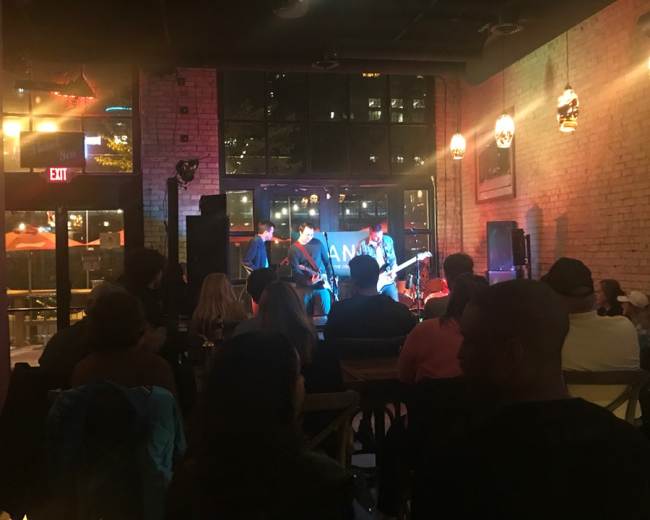 A small crowd watches a live band perform inside a dimly lit venue with large windows.