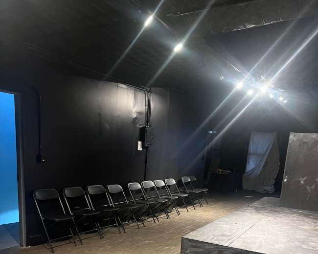 A black-walled theater space features a small stage with steps, several folding chairs arranged in a row, and a side door leading to another room.