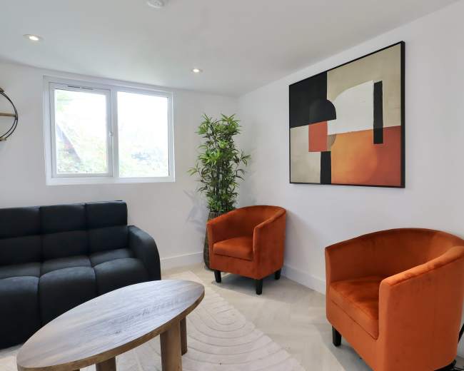 The image shows a modern living room with a black sectional sofa, two orange armchairs, a wooden coffee table, a decorative plant, and abstract wall art.