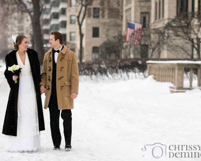 A couple dressed in wedding attire walks hand in hand through a snowy street lined with trees and buildings.