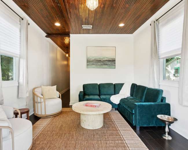 The image shows a bright, modern living room with a green sectional sofa, a round coffee table, and light-colored walls, accented by natural light from the windows.