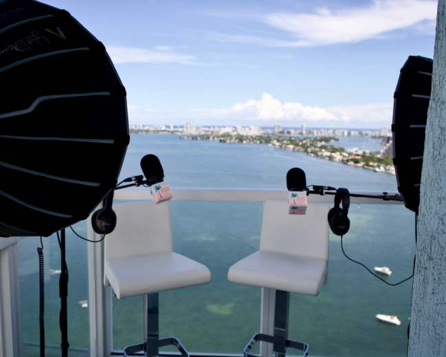 The scene shows two white barstools and microphones set up on a balcony overlooking a body of water and a city skyline in the distance.