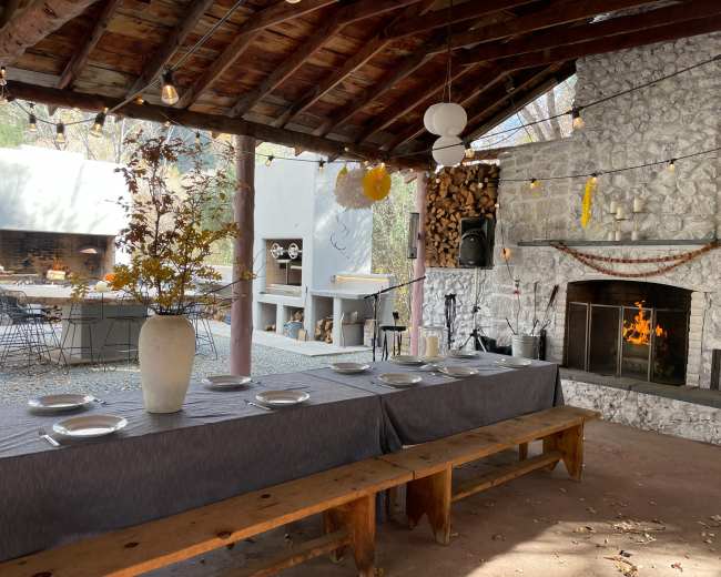 A rustic outdoor dining area under a wooden roof, featuring a long table with plates, a stone fireplace, and a gathering space decorated with lights and decorations.