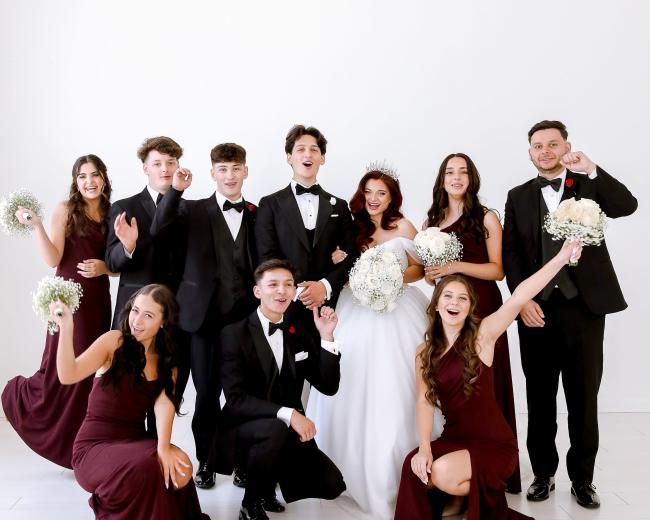 A group of young people dressed in formal attire poses together, holding bouquets and smiling for a photo in a bright, light-colored setting.