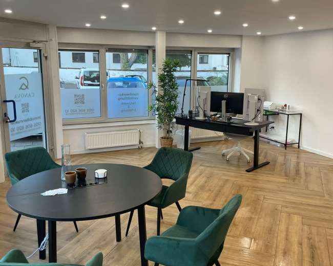 The image shows a modern office space with a round table surrounded by green chairs, a desk with two computer monitors, and large windows allowing natural light.