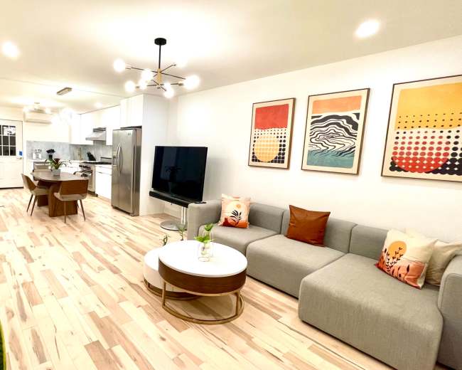 The image shows a modern living room with a gray sectional sofa, a round coffee table, a large TV, and a dining area, all featuring light-colored wood flooring and vibrant artwork on the walls.