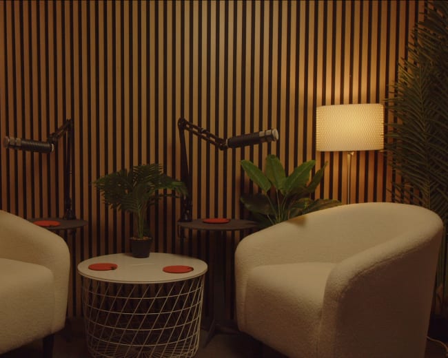 Two white armchairs are positioned around a small round table, set against a wooden slatted wall, with lamps and potted plants on either side.