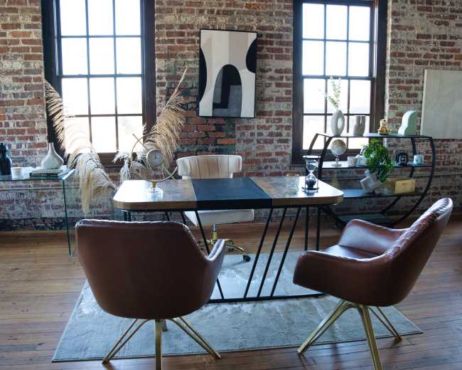 A modern office space features a wooden desk with a black mat, two brown chairs, large windows, and exposed brick walls adorned with artwork and decorative items.