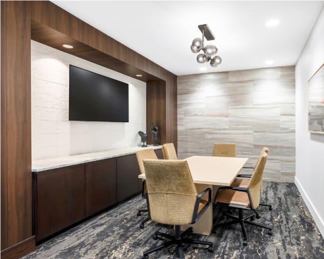 A conference room features a rectangular table surrounded by four chairs, with a large wall-mounted television and decorative wall paneling.