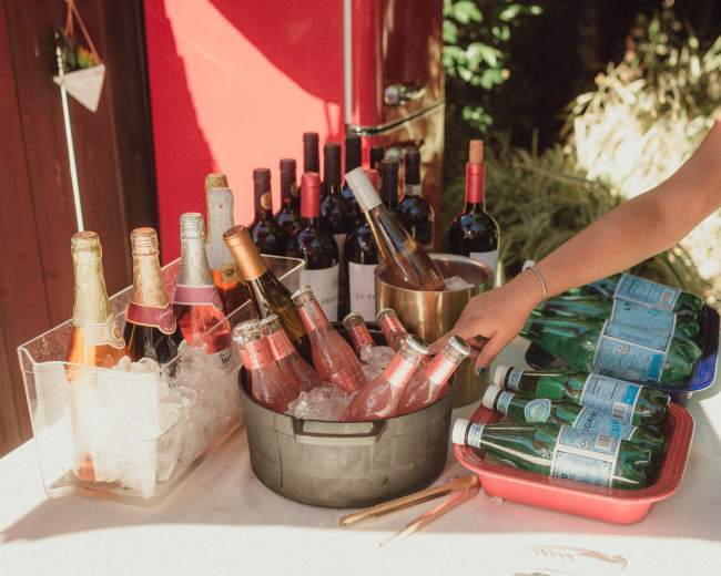 A variety of wine and sparkling water bottles are displayed in ice and on a table, with a person reaching for a drink.