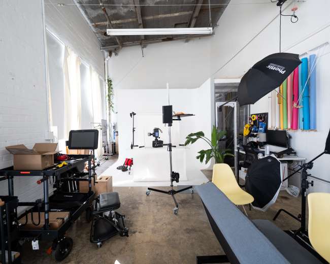 The image shows a photography studio with various equipment, including lights, cameras, and a backdrop, along with seating and organization carts.