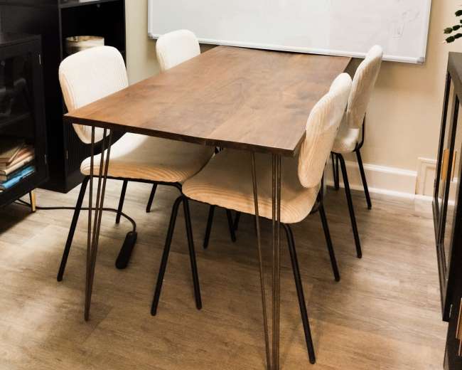 The image shows a simple dining table made of wood with five chairs arranged around it, in a room featuring a blank whiteboard on the wall.