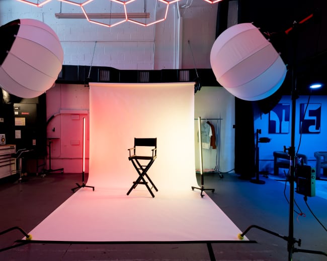 A photography studio is set up with a blank backdrop and a director's chair, illuminated by large softbox lights and neon accents.