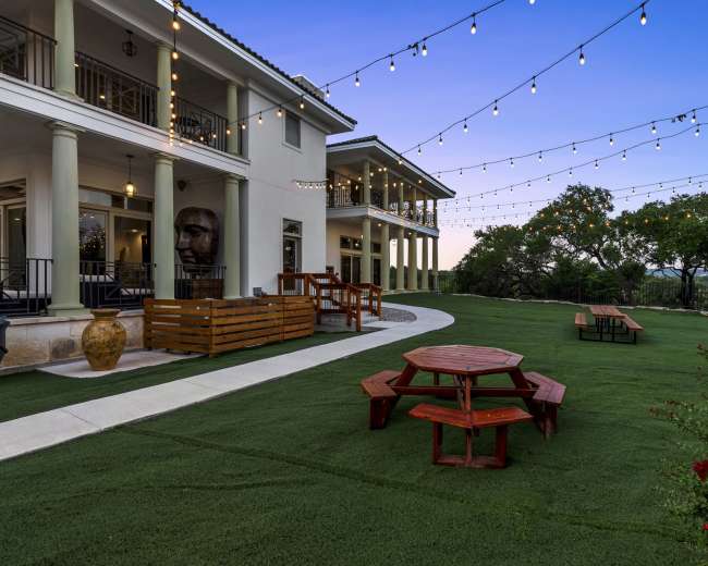 The scene features an outdoor area with string lights overhead, a red picnic table, and a landscaped lawn beside a modern building.