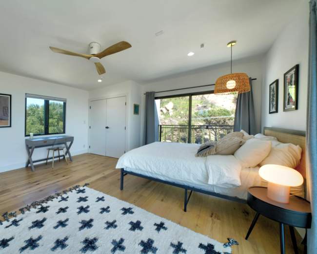 The image shows a spacious, modern bedroom featuring a bed, a desk with a chair, and large windows overlooking greenery.
