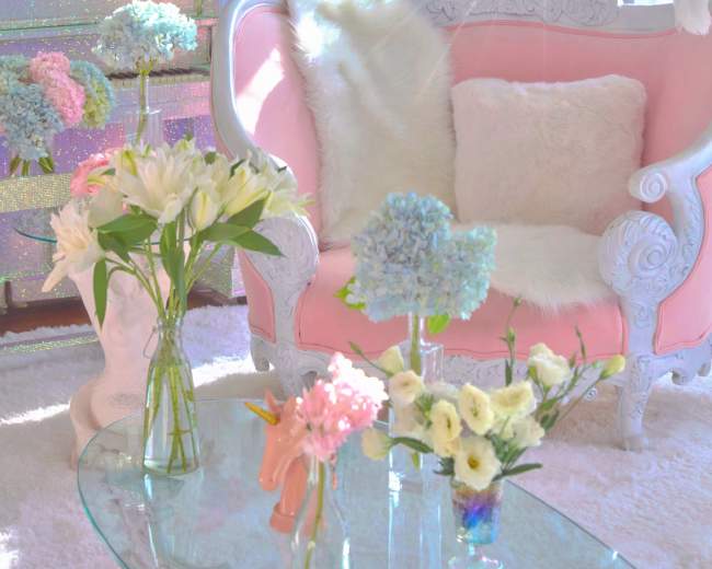 The scene features a light-filled room adorned with a pink sofa, floral arrangements in various vases, and a large disco ball reflecting light.