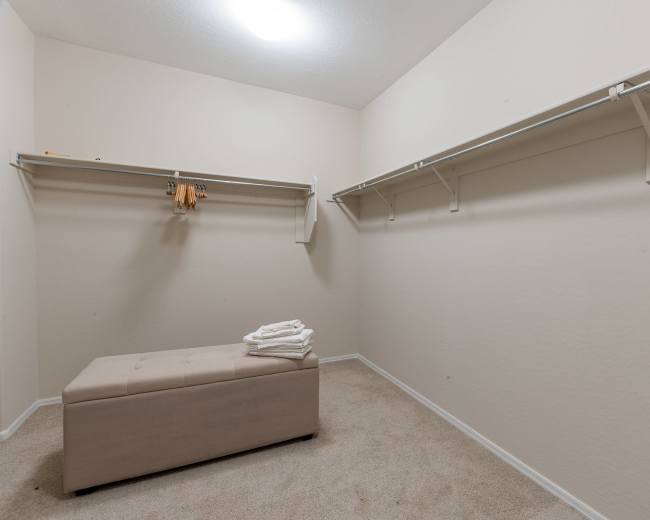 The image shows a blank room with beige walls, a carpeted floor, a storage bench in the center, and shelving along two walls.