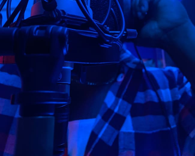 A person stands close to a microphone in a dimly lit room, with blue lighting illuminating the scene.