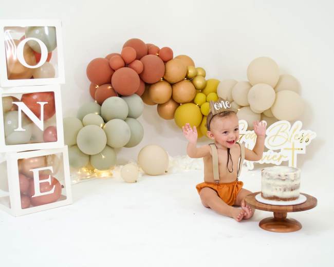 A baby wearing a party hat sits beside a decorated cake on a wooden stand, with balloon arrangements and a sign that reads "God Bless" in the background.