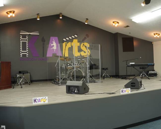 A stage set up for a performance, featuring a drum kit, keyboard, and sound equipment, with a large "KI Arts" sign on the wall behind it.