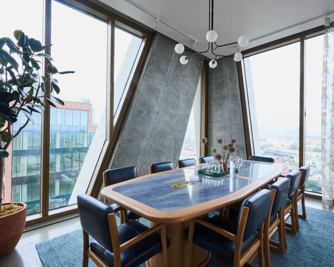 A modern dining room features a large wooden table surrounded by blue chairs, with large windows showcasing an urban view.