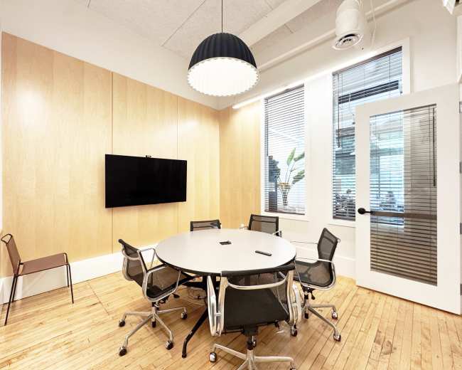 A modern conference room features a round table surrounded by six chairs, a large television mounted on the wall, and natural light from windows with blinds.