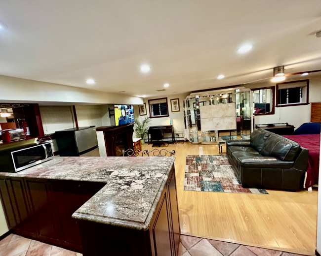 A well-lit basement living space featuring a kitchen area with a granite counter, a seating area with a dark sofa, and a sectioned-off sleeping area visible in the background.