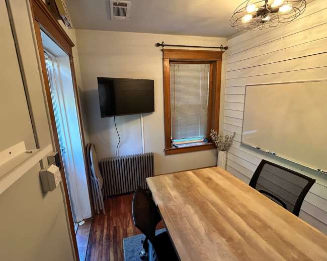 The image shows a small room with a wooden table and chairs, a TV mounted on the wall, and a whiteboard next to a window.