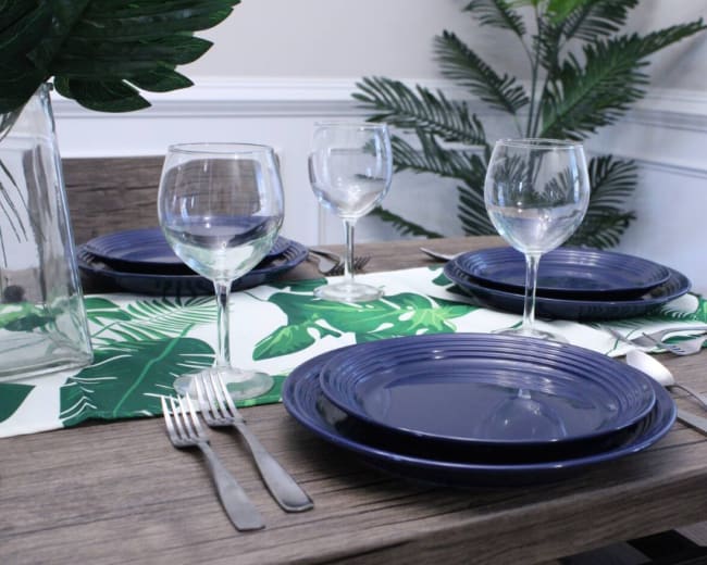 A dining table is set with navy plates, clear wine glasses, and silver cutlery, accompanied by a green leaf-patterned table runner and potted plants in the background.