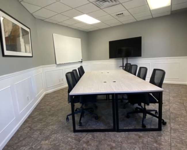 The image shows a conference room equipped with a long table surrounded by black chairs, a whiteboard on one wall, and a television mounted on another wall.