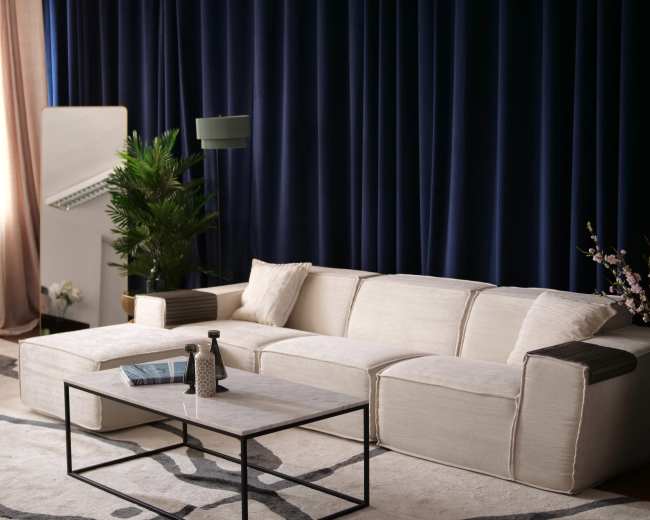 A modern living room features a light-colored sectional sofa, a coffee table, and a tall plant against dark blue curtains.