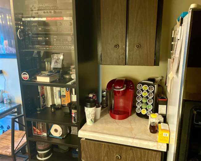 The image shows a kitchen corner featuring a coffee maker, a spice rack, and a shelving unit with various media equipment and books.