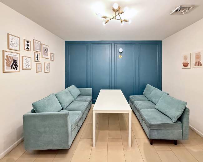 The scene features a modern meeting room with two light blue couches flanking a white rectangular table against a wall painted in a deep teal color, adorned with framed artwork.