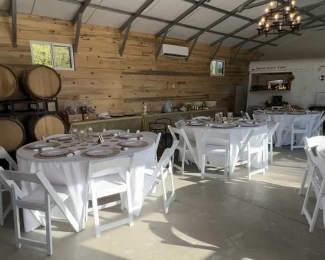 A rustic dining space with multiple round tables set with white tablecloths, surrounded by wooden walls and wine barrels.