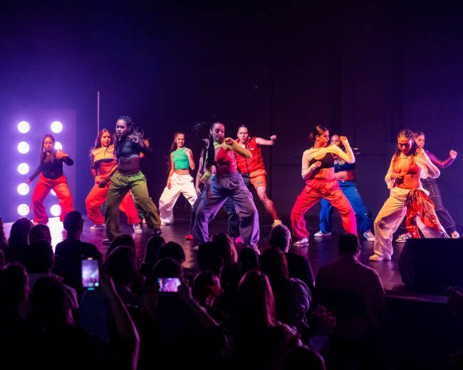 A group of dancers in colorful outfits performs on stage in front of an audience.