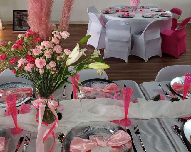 A dining setup features pink floral arrangements and table settings with silver plates and pink glassware, alongside a round table decorated in complementary colors.