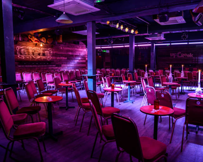 The image shows a dimly lit event space set up with rows of chairs and small tables, designed for an audience.