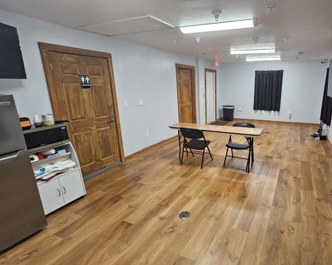 The image shows a sparsely furnished room with wooden flooring, featuring a table and two chairs, a refrigerator, and several flat-screen monitors mounted on the walls.