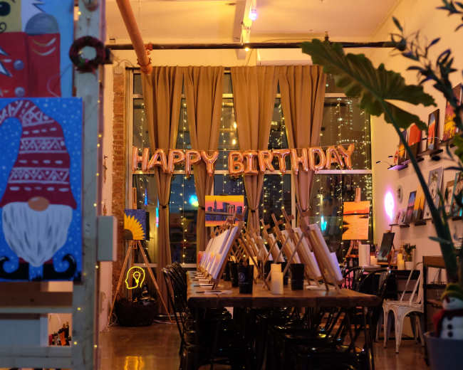 The image shows an art studio decorated for a birthday celebration, featuring a "HAPPY BIRTHDAY" sign, artwork on easels, and ambient lighting.
