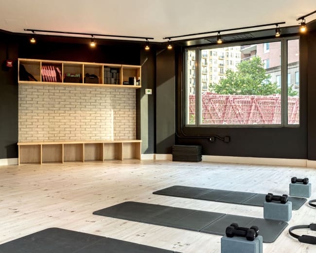 The image shows a modern fitness studio with large windows, wooden flooring, exercise mats, and weights neatly arranged on the floor.