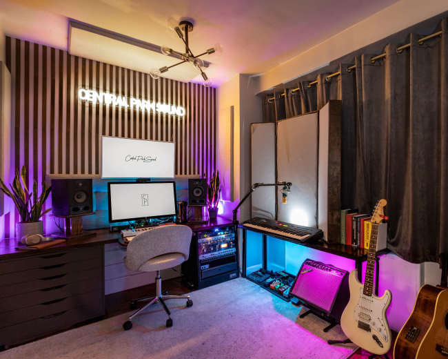The image shows a music studio with a desk featuring a computer setup, musical instruments including guitars, and colorful lighting.